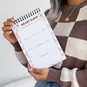 May include: A spiral-bound notepad with the words "BRAIN DUMP" at the top. The notepad has a floral design and sections for notes, including "To Do List," "To Call," "To Text," and "New Ideas." The notepad is held by a person wearing a brown and white sweater.