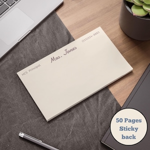 May include: A cream-colored notepad with the text "Mrs. Jones" and "HCS Principal" printed on it. The notepad has a sticky back and contains 50 pages. A phone number is also printed on the notepad. The notepad is on a wooden desk.