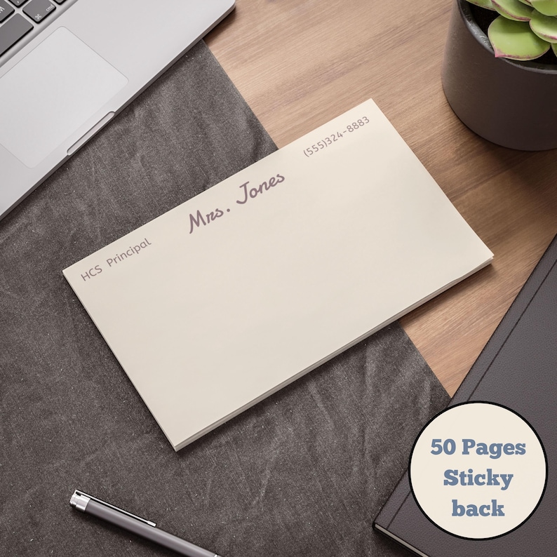May include: A cream-colored notepad with the text "Mrs. Jones" and "HCS Principal" printed on it. The notepad has a sticky back and contains 50 pages. A phone number is also printed on the notepad. The notepad is on a wooden desk.