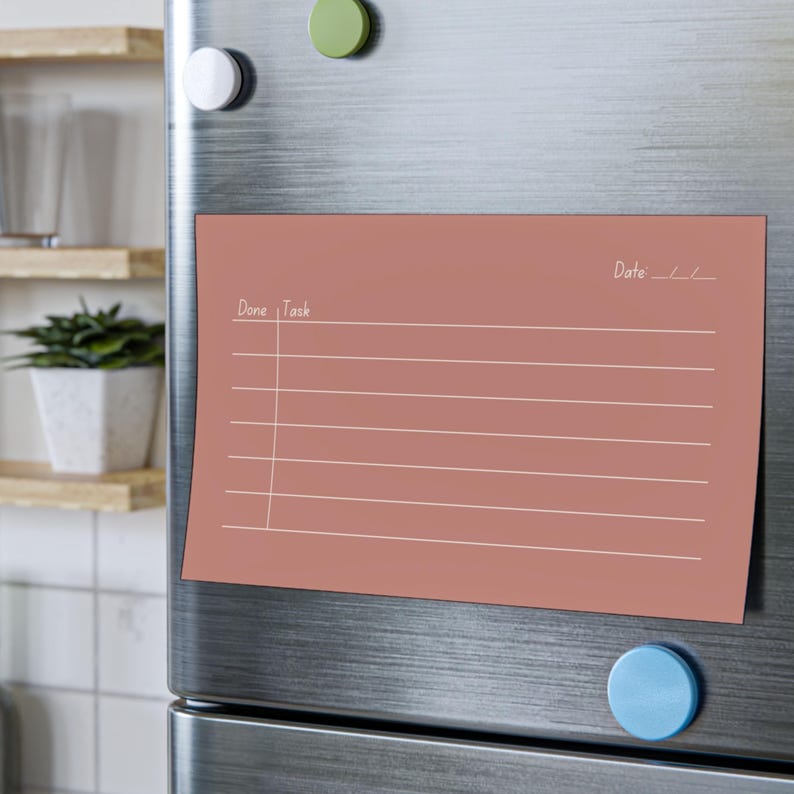 May include: A rectangular, dusty rose-colored magnetic notepad with white lines and text. The notepad is attached to a stainless steel refrigerator with magnets. The top of the notepad reads "Date: __/ __" and "Done | Task".
