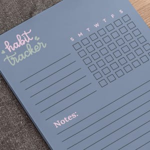 May include: A blue habit tracker notepad with a weekly calendar grid and space for notes. The words "habit tracker" are written in pink and green script. The notepad has horizontal lines for writing.