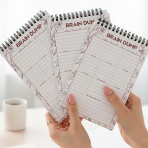 May include: Three spiral-bound notepads with the words "BRAIN DUMP" in burgundy. Each notepad has lined pages with sections for "TO CALL," "TO TEXT," and "NEW IDEAS." Floral accents frame the top of the pages. The notepads are held in a person's hands.