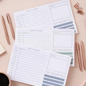 May include: A flat lay of daily plan notepads, pens, and office supplies on a light pink surface. The notepads have sections for schedule, notes, and top priorities. The pens and paperclips are rose gold. A cup of coffee is in the lower left corner.