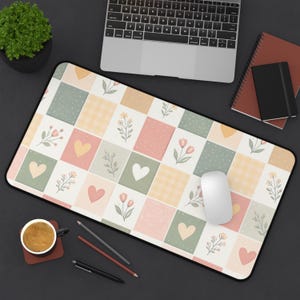 May include: A rectangular mousepad with a patchwork design featuring squares of pastel colors, hearts, flowers, and gingham patterns. A white mouse rests on the pad. A laptop, notebooks, pens, and a cup of coffee are also visible.