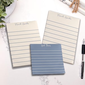 May include: Three notepads with horizontal lines. Two are off-white with "Maxwell Associates" printed at the top. The third is blue with "Heidi Thomas" printed at the top. A black pen is on the right.