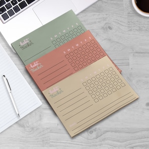May include: Three pastel-colored habit tracker notepads. Each notepad features a calendar grid for tracking daily habits, with the days of the week labeled. The phrase "habit tracker" is written in a script font. The notepads are stacked on a wooden surface.
