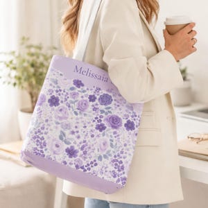 May include: A lavender tote bag with a floral pattern featuring purple roses and other flowers. The bag has a white strap and the name "Melissaia" printed on the top. The bag is being carried by a person wearing a white blazer.