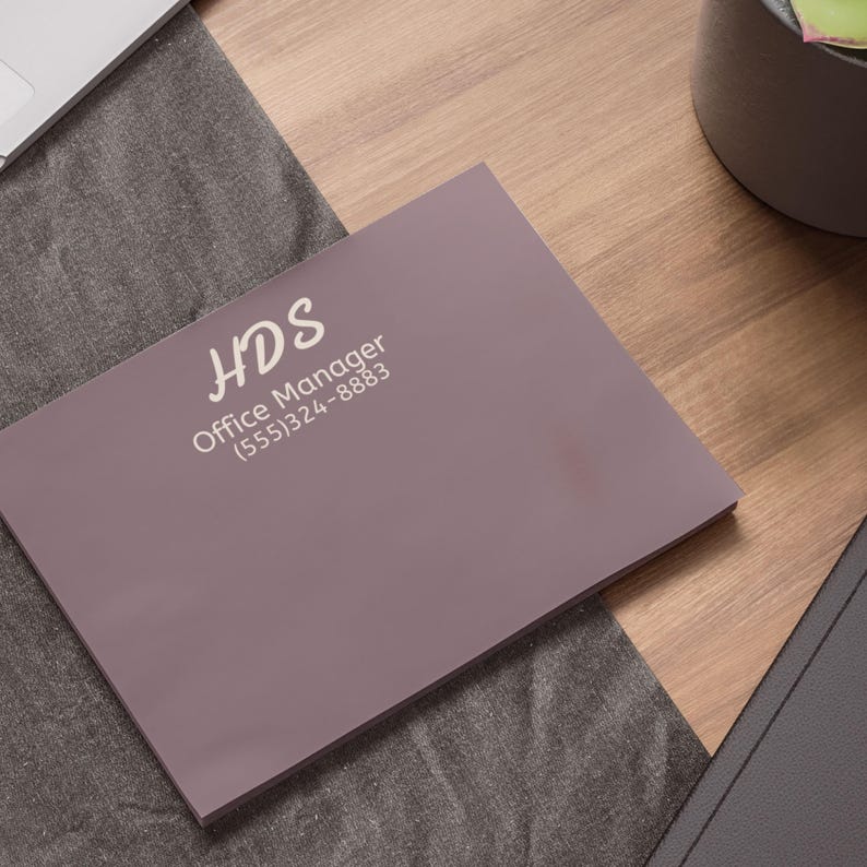 May include: A mauve-colored notepad with the text "HDS Office Manager (555)324-8883" printed in white. The notepad is on a wooden surface next to a gray cloth and a laptop.