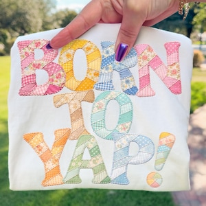 Könnte beinhalten: Weißes T-Shirt mit dem Schriftzug "BORN TO YAP!" in Patchwork-Buchstaben. Die Buchstaben bestehen aus verschiedenen pastellfarbenen Stoffen mit Blumen- und Vichy-Mustern. Ein farbenfrohes Ausrufezeichen und ein Regenbogenkreis vervollständigen das Design.