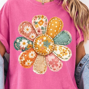 May include: A bright pink t-shirt featuring a large, colorful flower design. The flower is made of various fabric patches with floral and polka dot patterns, stitched together. The shirt has rolled-up sleeves.