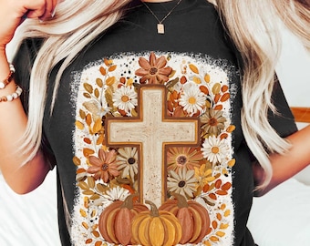 May include: Black t-shirt featuring a cross design with pumpkins and flowers. The cross is surrounded by fall-colored flowers and leaves. Three pumpkins are at the bottom. The design has a distressed, vintage look.
