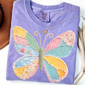 Op de afbeelding: Een lavendelkleurig Comfort Colors T-shirt met een patchwork vlinderontwerp in pasteltinten. Een gouden ketting met een kleine hanger ligt op het shirt. De vlindervleugels zijn gemaakt van bloemen-, ruit- en effen stoffen.