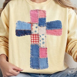 以下が含まれることがあります： パッチワークの十字架のデザインが施された、薄い黄色のスウェットシャツ。十字架は、デニム、ピンク、ブルー、花柄の生地の正方形で作られています。スウェットシャツはクルーネックで長袖です。