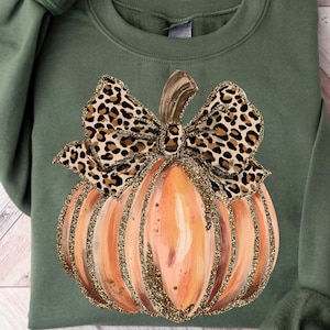 May include: Green sweatshirt featuring a large pumpkin design. The pumpkin is orange with gold glitter accents and a leopard print bow. The design is centered on the front of the sweatshirt, creating a festive fall theme.