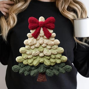 May include: Black sweatshirt featuring a textured Christmas tree design. The tree is composed of layers of green, beige, and brown, topped with a red bow. The design is a festive and cozy holiday-themed apparel.