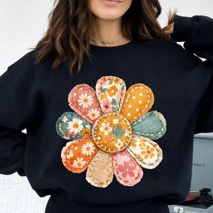 Może przedstawiać: Czarna bluza z dużym, kolorowym wzorem kwiatowym. Płatki kwiatu są w stylu patchwork, z r&oacute;żnymi wzorami kwiatowymi i w kropki w odcieniach pomarańczu, r&oacute;żu, zieleni i ż&oacute;łci. Środek kwiatu jest w jednolitym pomarańczowym kolorze.