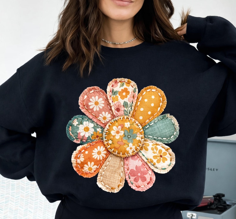 Może przedstawiać: Czarna bluza z dużym, kolorowym wzorem kwiatowym. Płatki kwiatu są w stylu patchwork, z r&oacute;żnymi wzorami kwiatowymi i w kropki w odcieniach pomarańczu, r&oacute;żu, zieleni i ż&oacute;łci. Środek kwiatu jest w jednolitym pomarańczowym kolorze.