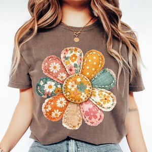 Może przedstawiać: Brązowa koszulka z dużym, kolorowym wzorem kwiatowym. Kwiat wykonany jest z r&oacute;żnych łat materiału w odcieniach pomarańczu, r&oacute;żu, ż&oacute;łci i zieleni, z kwiatowymi i groszkowymi wzorami. Wz&oacute;r jest wyśrodkowany na koszulce.