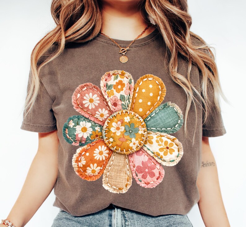 Może przedstawiać: Brązowa koszulka z dużym, kolorowym wzorem kwiatowym. Kwiat wykonany jest z r&oacute;żnych łat materiału w odcieniach pomarańczu, r&oacute;żu, ż&oacute;łci i zieleni, z kwiatowymi i groszkowymi wzorami. Wz&oacute;r jest wyśrodkowany na koszulce.