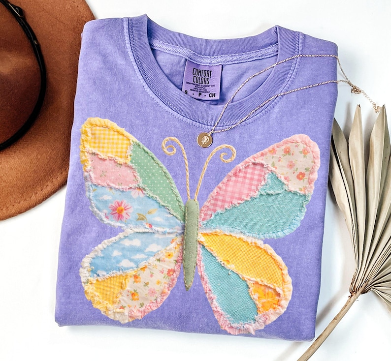 May include: A lavender Comfort Colors t-shirt featuring a patchwork butterfly design in various pastel colors. A gold necklace with a small pendant rests on the shirt. The butterfly has wings made of floral, gingham, and solid fabrics.