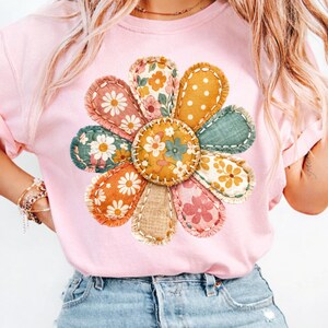 Może przedstawiać: Jasnor&oacute;żowa koszulka z dużym, kolorowym wzorem kwiatowym. Kwiat ma płatki z tkaniny w r&oacute;żnych wzorach i kolorach, w tym nadruki kwiatowe i kropki. Projekt jest wyśrodkowany na koszulce.