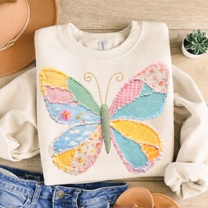 May include: Cream-colored sweatshirt featuring a patchwork butterfly design. The butterfly wings are composed of various fabric patterns in pastel colors, including pink, blue, yellow, and green. The sweatshirt is paired with denim shorts and sandals.