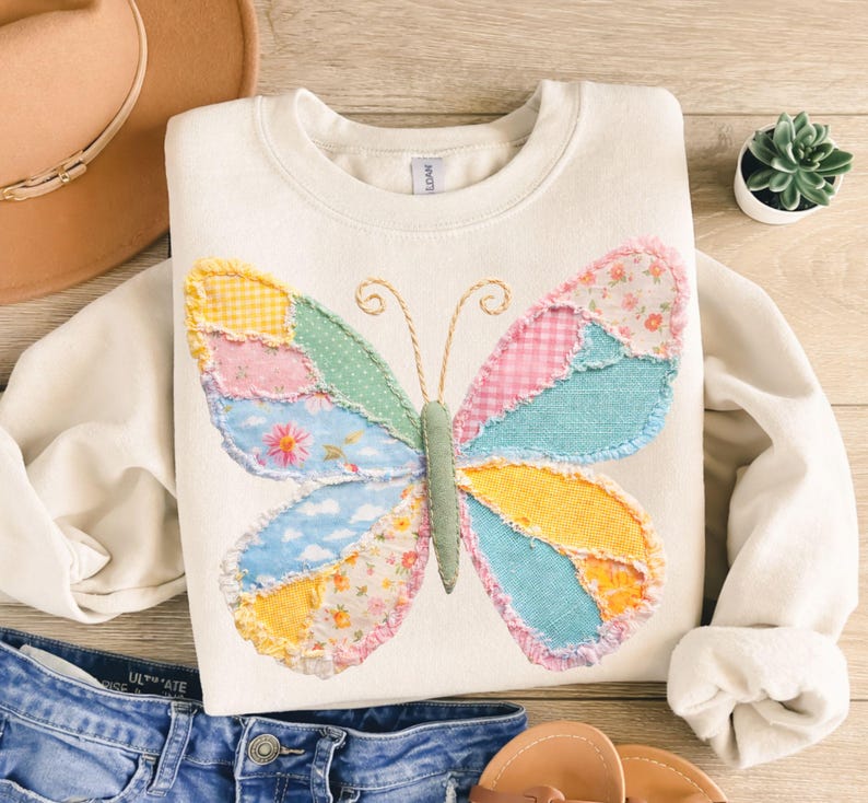 May include: Cream-colored sweatshirt featuring a patchwork butterfly design. The butterfly wings are composed of various fabric patterns in pastel colors, including pink, blue, yellow, and green. The sweatshirt is paired with denim shorts and sandals.
