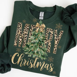 K&ouml;nnte beinhalten: Ein dunkelgr&uuml;nes Sweatshirt mit den Worten "MERRY Christmas" in Goldglitzer und Leopardenmuster. Ein Weihnachtsbaum mit einer goldenen Schleife befindet sich in der Mitte. Goldene Schneeflocken sind um den Text herum verstreut.