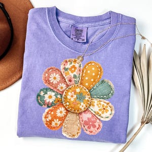 Może przedstawiać: Jasnofioletowa koszulka Comfort Colors z dużą, kolorową aplikacją kwiatową. Kwiat ma płatki w r&oacute;żnych wzorach w kolorze pomarańczowym, r&oacute;żowym, zielonym i ż&oacute;łtym. Złoty naszyjnik z wisiorkiem spoczywa na koszulce. Koszulka ma małą metkę z napisem "Comfort Colors".