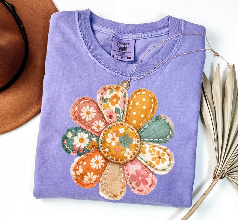 Może przedstawiać: Jasnofioletowa koszulka Comfort Colors z dużą, kolorową aplikacją kwiatową. Kwiat ma płatki w r&oacute;żnych wzorach w kolorze pomarańczowym, r&oacute;żowym, zielonym i ż&oacute;łtym. Złoty naszyjnik z wisiorkiem spoczywa na koszulce. Koszulka ma małą metkę z napisem "Comfort Colors".