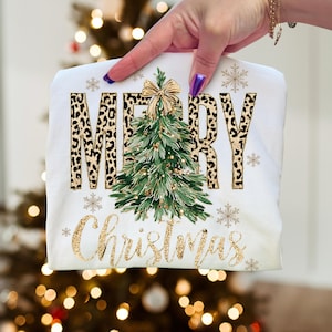 K&ouml;nnte beinhalten: Wei&szlig;es T-Shirt mit Weihnachtsdesign. Das Wort "MERRY" ist im Leopardenmuster und "Christmas" in goldfarbenem Glitzerschriftzug gehalten. Ein gr&uuml;ner Weihnachtsbaum mit goldener Schleife befindet sich in der Mitte, mit goldenen Schneeflocken.