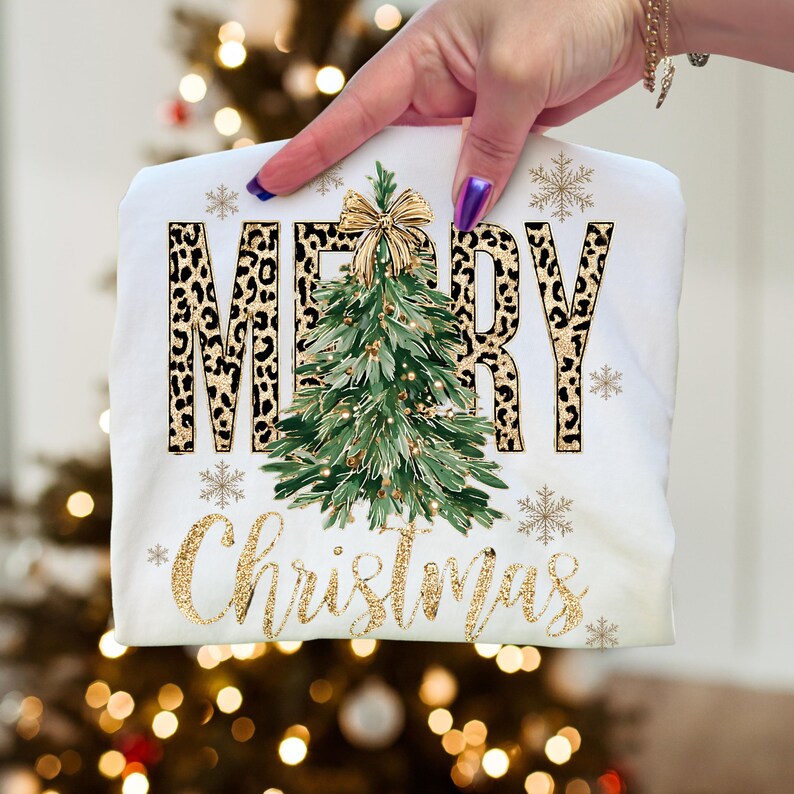 K&ouml;nnte beinhalten: Wei&szlig;es T-Shirt mit Weihnachtsdesign. Das Wort "MERRY" ist im Leopardenmuster und "Christmas" in goldfarbenem Glitzerschriftzug gehalten. Ein gr&uuml;ner Weihnachtsbaum mit goldener Schleife befindet sich in der Mitte, mit goldenen Schneeflocken.