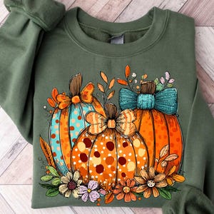 May include: Green sweatshirt featuring a colorful pumpkin design. The pumpkins are orange with polka dots and bows, and surrounded by flowers and leaves. The design has a fall or autumn theme.