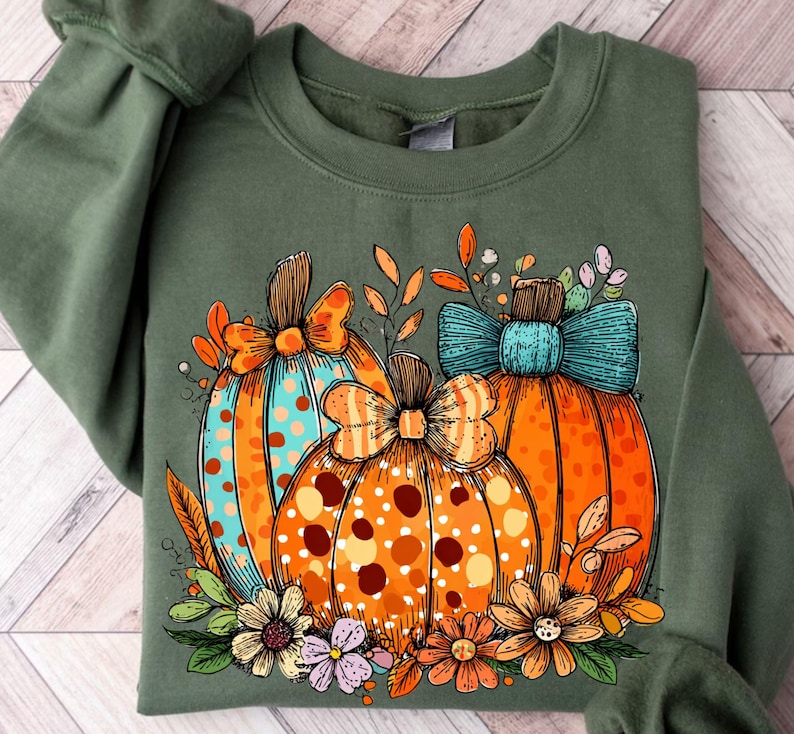 May include: Green sweatshirt featuring a colorful pumpkin design. The pumpkins are orange with polka dots and bows, and surrounded by flowers and leaves. The design has a fall or autumn theme.