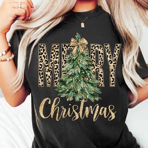 K&ouml;nnte beinhalten: Schwarzes T-Shirt mit einem Weihnachtsdesign. Das Wort "MERRY" ist im Leopardenmuster gehalten, und ein gr&uuml;ner Weihnachtsbaum mit goldener Schleife befindet sich in der Mitte. Die Worte "Christmas" sind in goldener Schrift geschrieben.