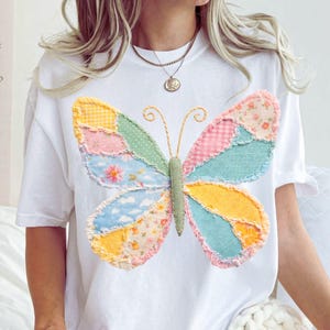 May include: White t-shirt featuring a colorful patchwork butterfly design. The butterfly wings are made of various patterned fabrics in pastel colors, including pink, blue, yellow, and green. The butterfly's body is green, and the antennae are gold.