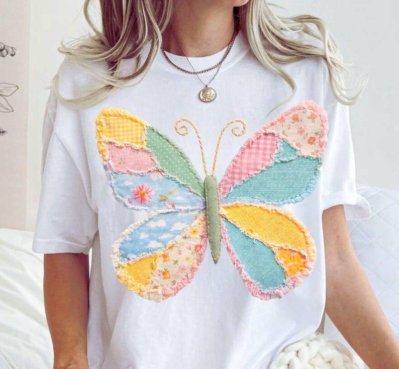 May include: White t-shirt featuring a colorful patchwork butterfly design. The butterfly wings are made of various patterned fabrics in pastel colors, including pink, blue, yellow, and green. The butterfly's body is green, and the antennae are gold.