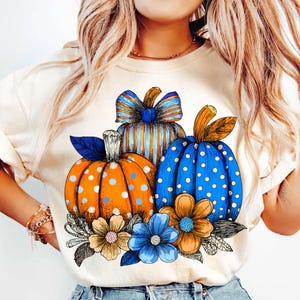 May include: Cream-colored t-shirt featuring a fall-themed graphic. The design includes three pumpkins in orange and blue with white polka dots and stripes, accented by blue and tan flowers and leaves. A festive autumn design.