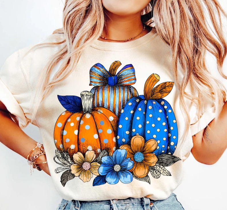 May include: Cream-colored t-shirt featuring a fall-themed graphic. The design includes three pumpkins in orange and blue with white polka dots and stripes, accented by blue and tan flowers and leaves. A festive autumn design.