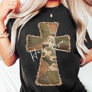 May include: Black t-shirt featuring a patchwork cross design with camouflage and burlap textures. The words "He is Risen" are written across the cross in a white script. The shirt has a casual, relaxed fit.