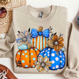 May include: Beige sweatshirt featuring a colorful pumpkin design. The pumpkins are orange, blue, and striped, adorned with flowers and bows. A fall-themed graphic for seasonal wear.