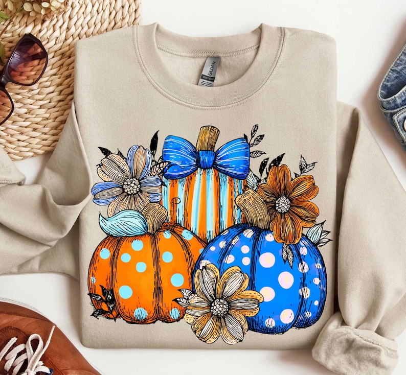 May include: Beige sweatshirt featuring a colorful pumpkin design. The pumpkins are orange, blue, and striped, adorned with flowers and bows. A fall-themed graphic for seasonal wear.
