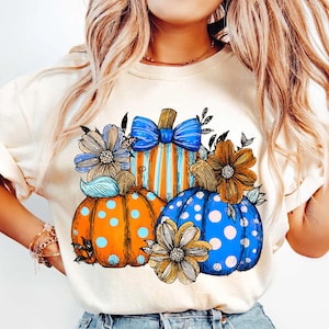 May include: Cream-colored t-shirt featuring a colorful pumpkin design. The pumpkins are orange, blue, and striped, adorned with blue bows and flowers. The design has a whimsical, hand-drawn style, perfect for autumn.