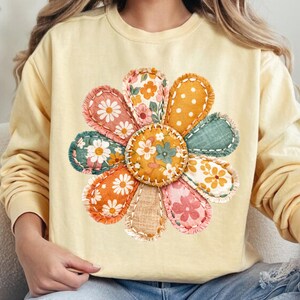 Może przedstawiać: Jasnoż&oacute;łta bluza z dużą, kolorową aplikacją kwiatową. Kwiat ma r&oacute;żne wzorzyste płatki w kolorze pomarańczowym, r&oacute;żowym, zielonym i białym, z przeszytym wzorem. Środek kwiatu to mniejsze koło z kwiatowymi detalami.