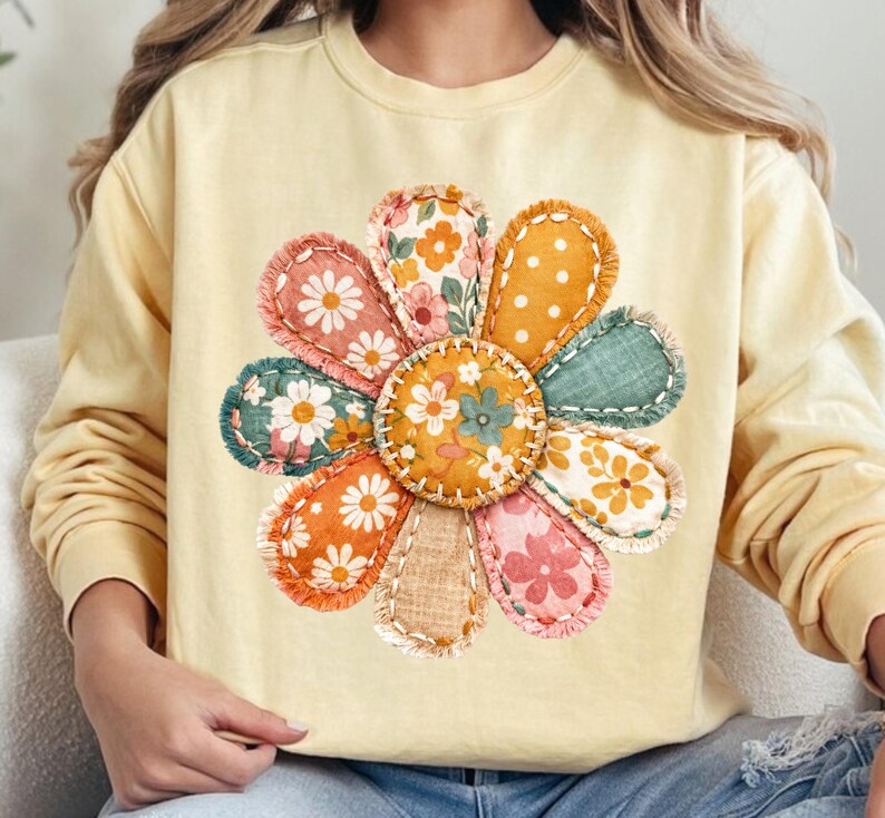 Może przedstawiać: Jasnoż&oacute;łta bluza z dużą, kolorową aplikacją kwiatową. Kwiat ma r&oacute;żne wzorzyste płatki w kolorze pomarańczowym, r&oacute;żowym, zielonym i białym, z przeszytym wzorem. Środek kwiatu to mniejsze koło z kwiatowymi detalami.