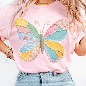 May include: A light pink t-shirt featuring a large, colorful butterfly applique. The butterfly has patchwork wings in various pastel patterns, including floral, gingham, and cloud designs. The butterfly's body is green, and the antennae are gold.