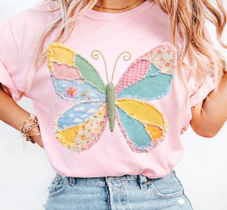 May include: A light pink t-shirt featuring a large, colorful butterfly applique. The butterfly has patchwork wings in various pastel patterns, including floral, gingham, and cloud designs. The butterfly's body is green, and the antennae are gold.