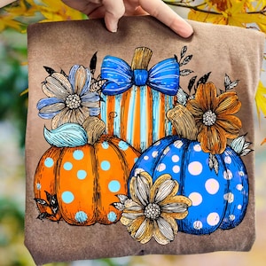 May include: Brown t-shirt featuring a colorful fall design. The design includes three pumpkins in orange and blue with floral accents. The central pumpkin has orange and blue stripes and a blue bow.