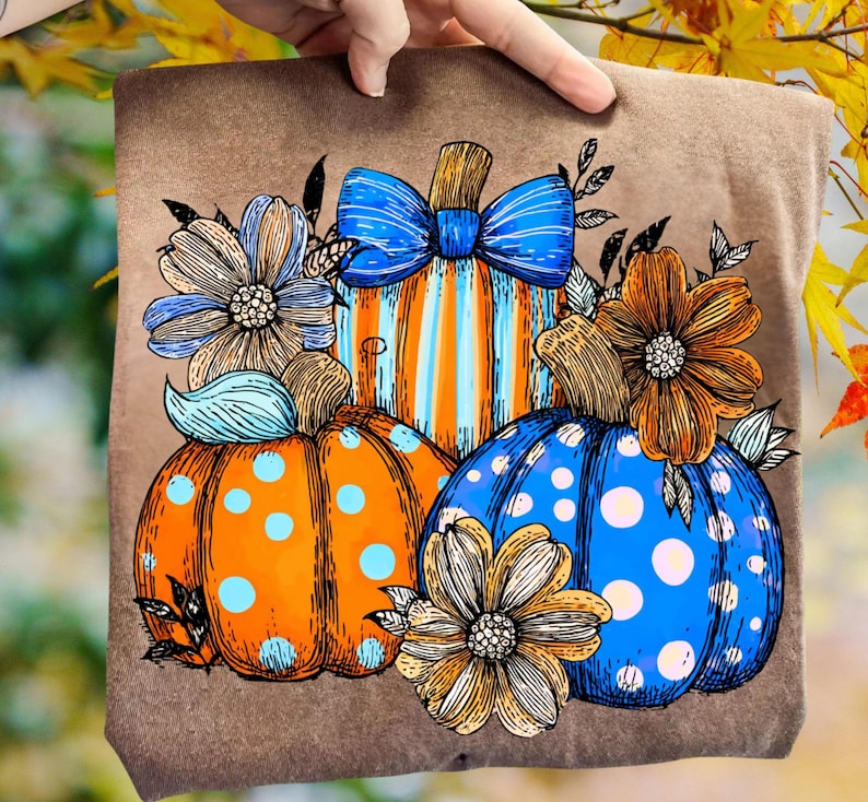May include: Brown t-shirt featuring a colorful fall design. The design includes three pumpkins in orange and blue with floral accents. The central pumpkin has orange and blue stripes and a blue bow.