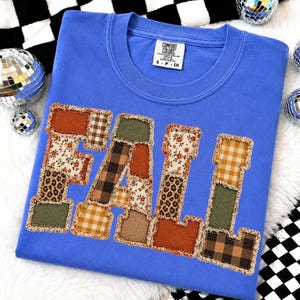 Puede incluir: Una camiseta azul de cuello redondo con la palabra FALL en grandes letras de tela con bordes deshilachados. Cada letra está construida con varios parches estampados, incluyendo flores, estampado de leopardo, cuadros, vichy y tela lisa en color oliva y naranja quemado.